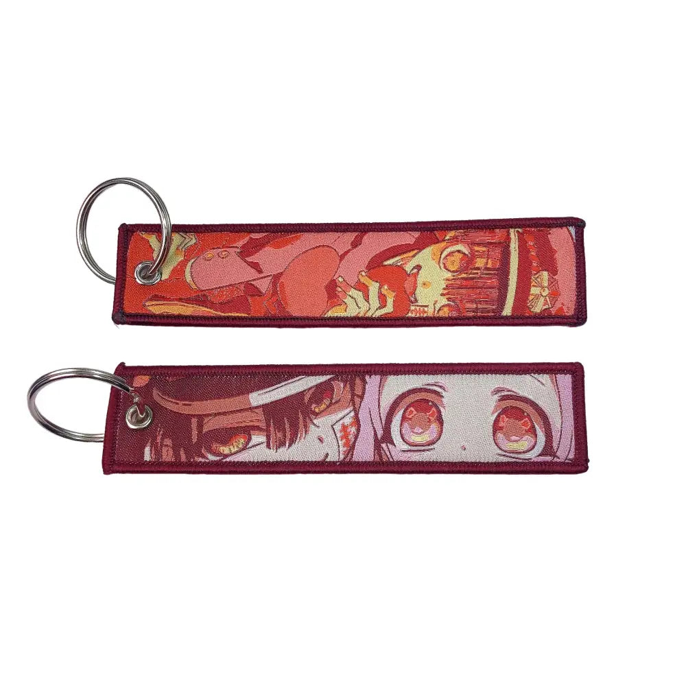 Two rectangular embroidered anime lock keychains with vibrant red and pink hues on dark maroon jet tag fabric base