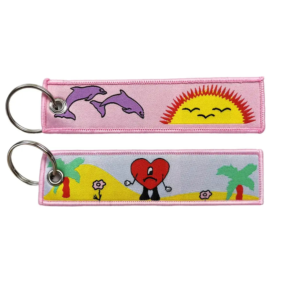 Pink embroidered key fob with colorful anime cartoon designs featuring purple dolphins, smiling sun, and sad heart key tag
