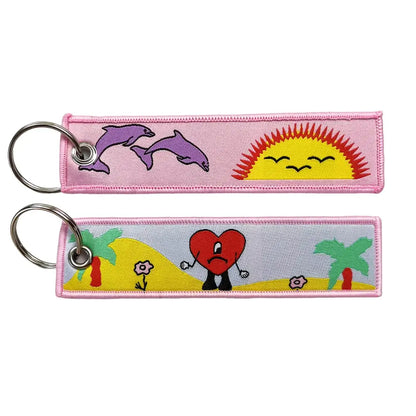 Pink embroidered key fob with colorful anime cartoon designs featuring purple dolphins, smiling sun, and sad heart key tag