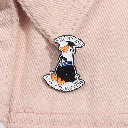 Anime character enamel pin of a black and orange duck wearing a graduation cap and gown with trust me im a ducktor text