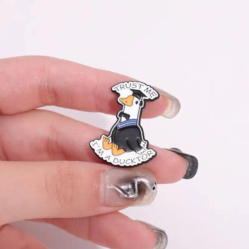 Black and white enamel pin featuring a cartoon duck with a graduation cap and trust me im a ducktor text metal brooch