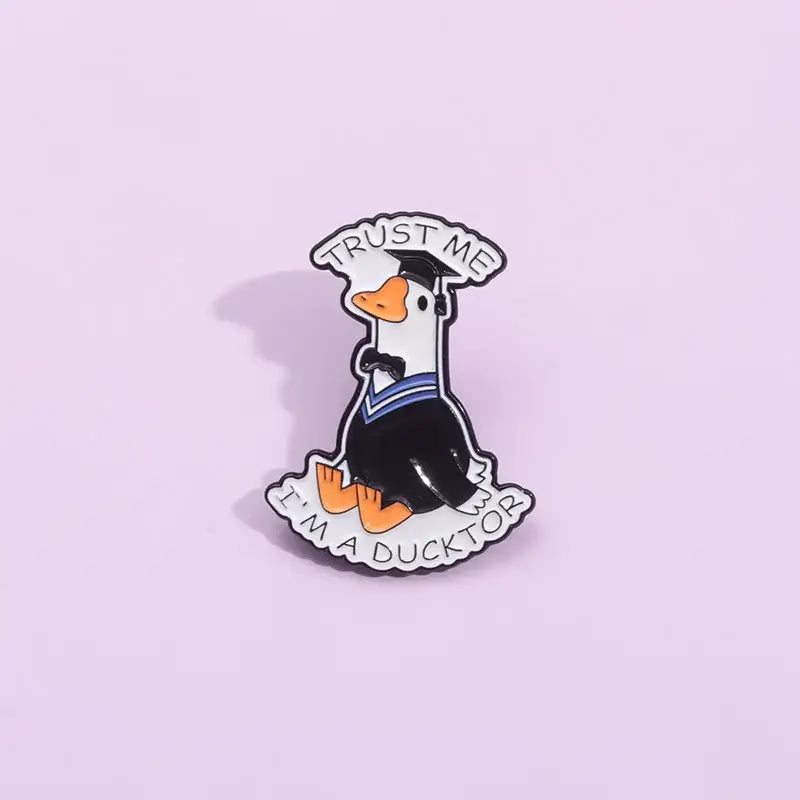Black and white enamel pin of a cartoon duck in graduation attire with orange beak and feet metal brooch