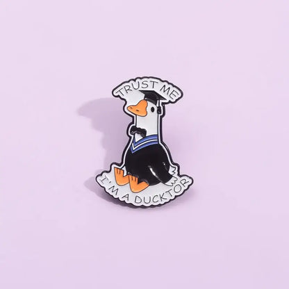 Black and white enamel pin of a cartoon duck in graduation attire with orange beak and feet metal brooch