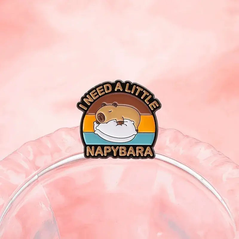 A charming anime enamel brooch of a sleeping capybara on a pillow with sunset gradient and napybara text