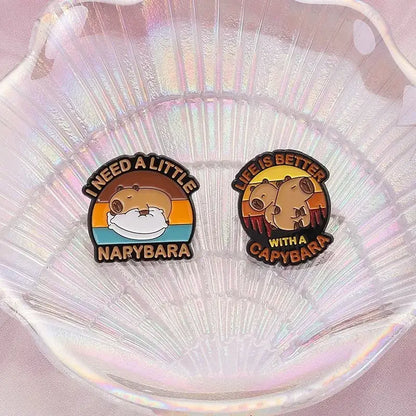 Two circular anime enamel brooch pins featuring cute capybara with colorful sunset backgrounds and text designs