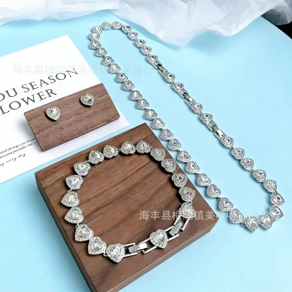 Heart-shaped crystal sterling silver tennis necklace and matching earrings displayed on wooden trays