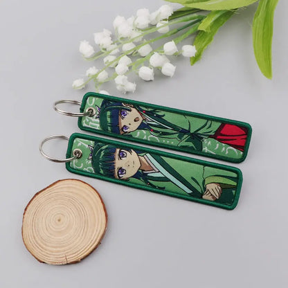 Green embroidered metal maomao key chain with anime characters purple eyes and green hair on white flower background key ring