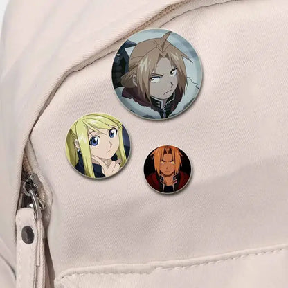 Three colorful fullmetal alchemist enamel brooch pins with handmade designs on light-colored fabric background