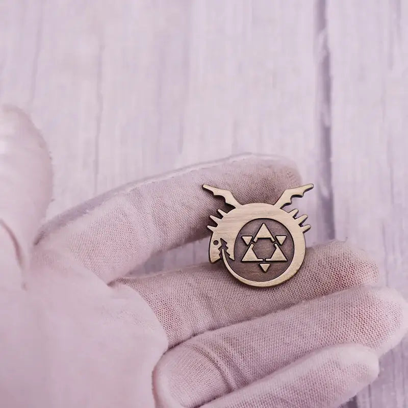 A light brown wooden fullmetal alchemist logo enamel pin with circular design, triangular patterns, and wing-like metal badge