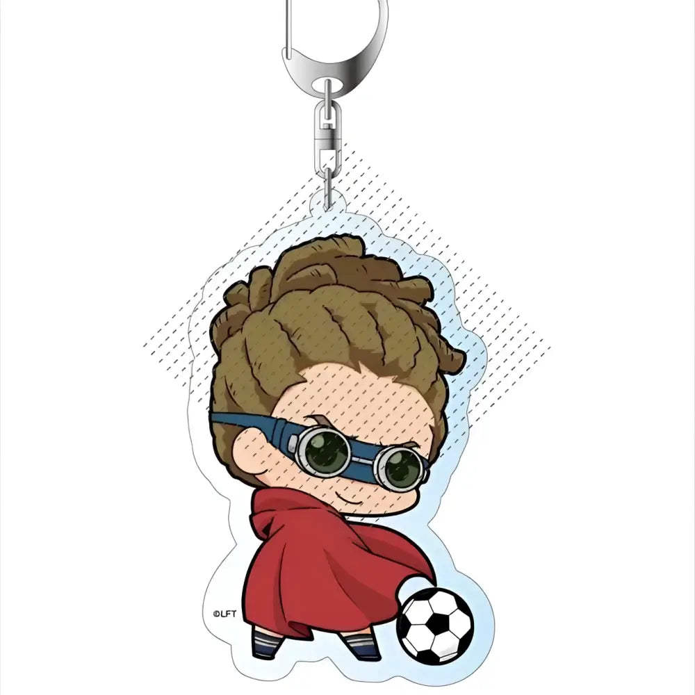 A clear acrylic keychain featuring a chibi style brown curly haired character with blue goggles and red cape holding a soccer ball