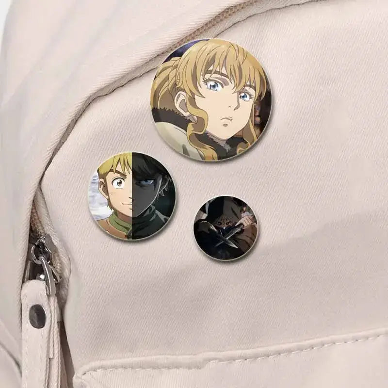 Three circular vinland saga thorfinn button brooch pins with detailed tinplate illustrations on light fabric surface
