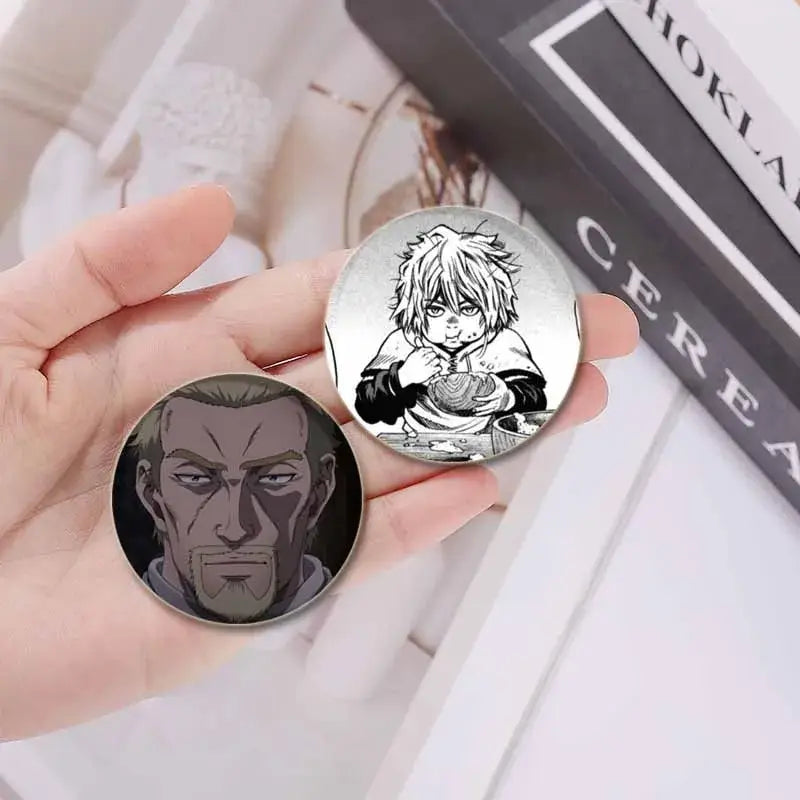 Two circular vinland saga thorfinn tinplate soft badges featuring a boy with spiky hair and an intense older man