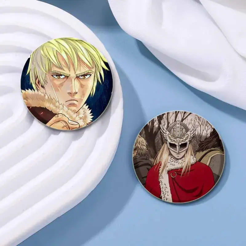Two circular vinland saga thorfinn button pins featuring a blonde figure with fur collar and a masked warrior in red cloak