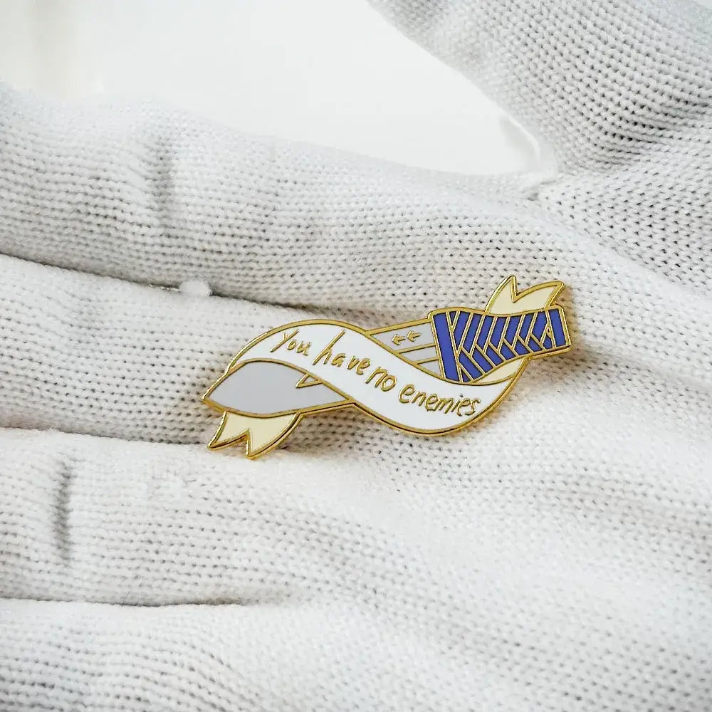 Gold-toned vinland saga thorfinn enamel pin with ribbon design saying you have no enemies and blue-striped emblem on white fabric