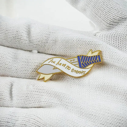 Gold-toned vinland saga thorfinn enamel pin with ribbon design saying you have no enemies and blue-striped emblem on white fabric