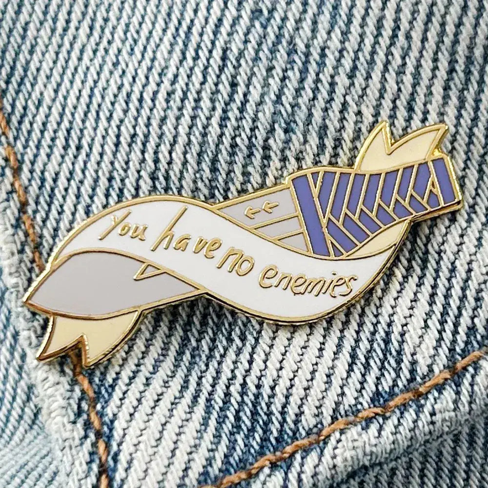 Gold-toned vinland saga thorfinn enamel pin shaped like a ribbon with purple white stripes and cursive phrase you have no enemies