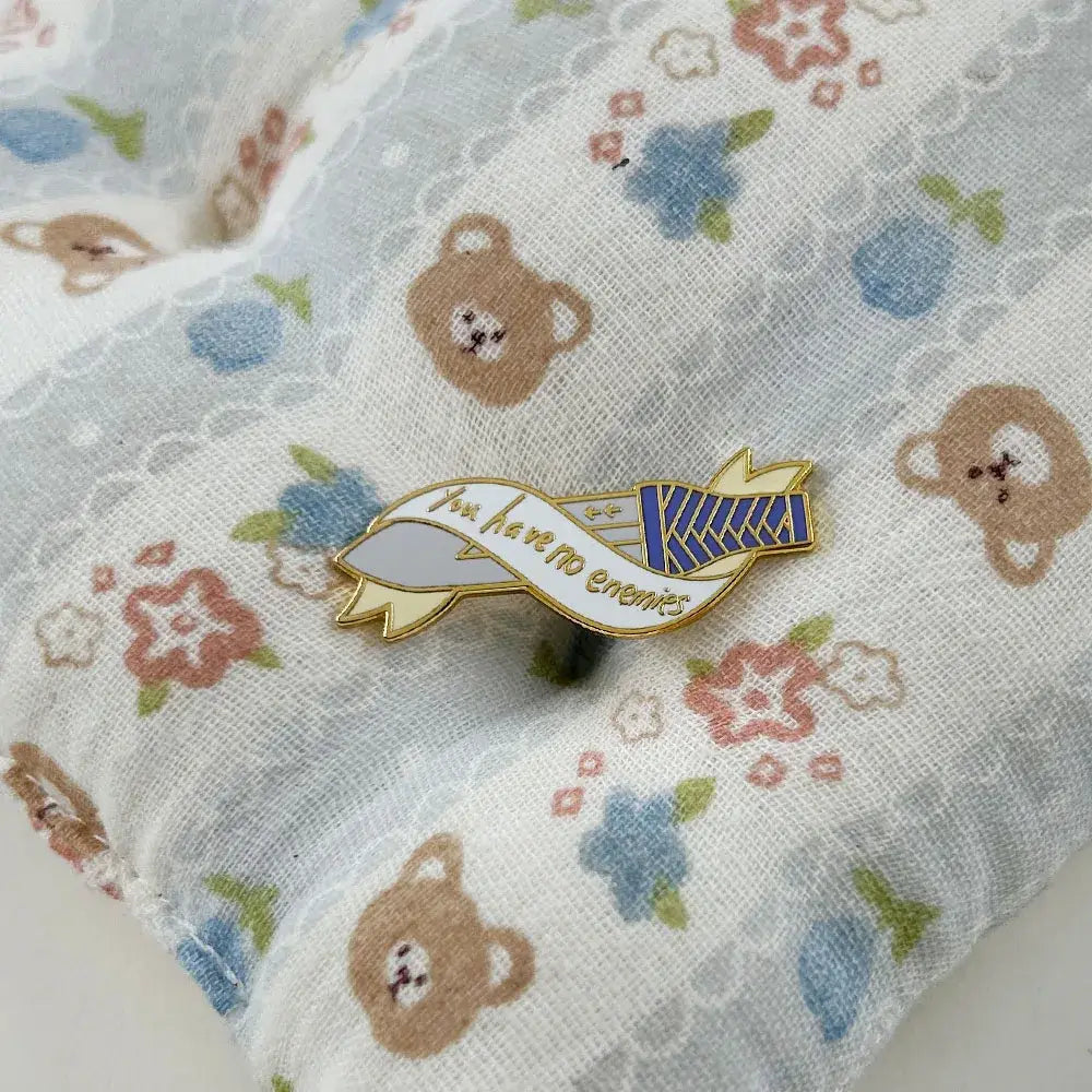 Gold-toned vinland saga thorfinn enamel pin shaped like a ribbon banner with you have no enemies text and blue white stripes